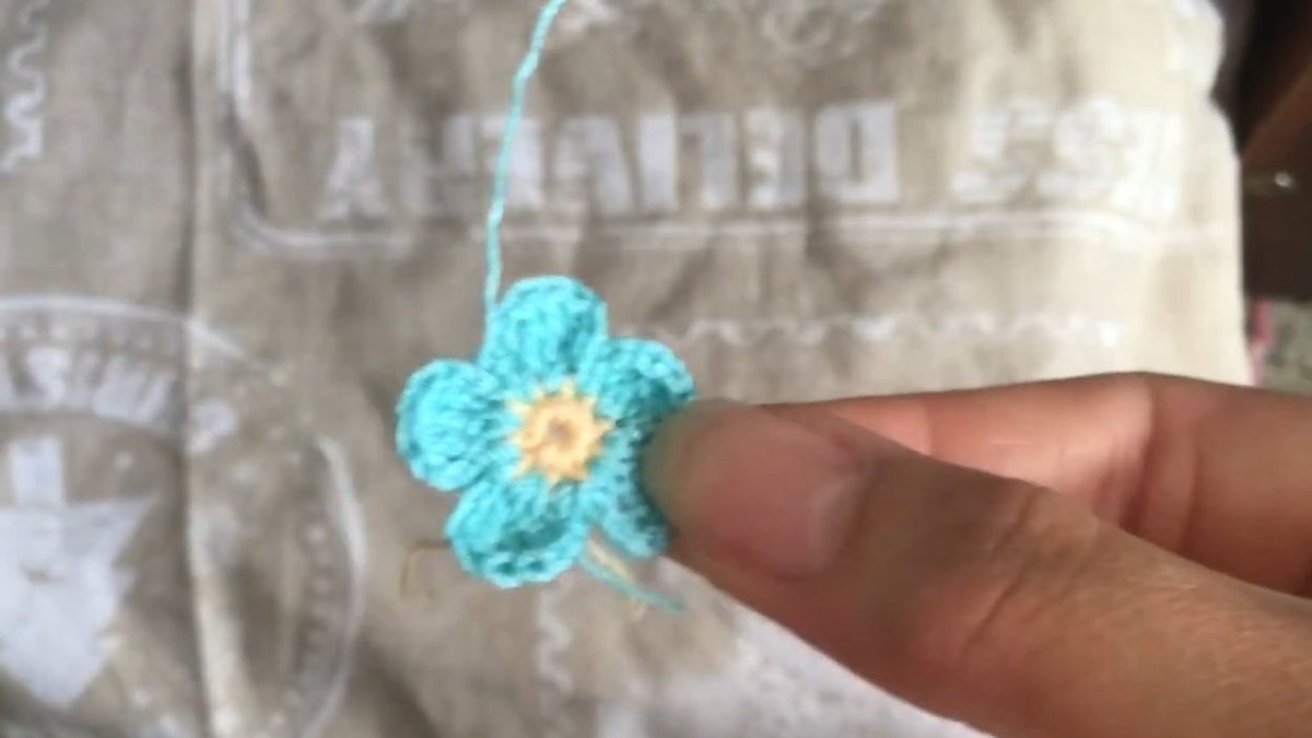 Finished teal crochet forget me not flower with a peach centre resting on a fingertip