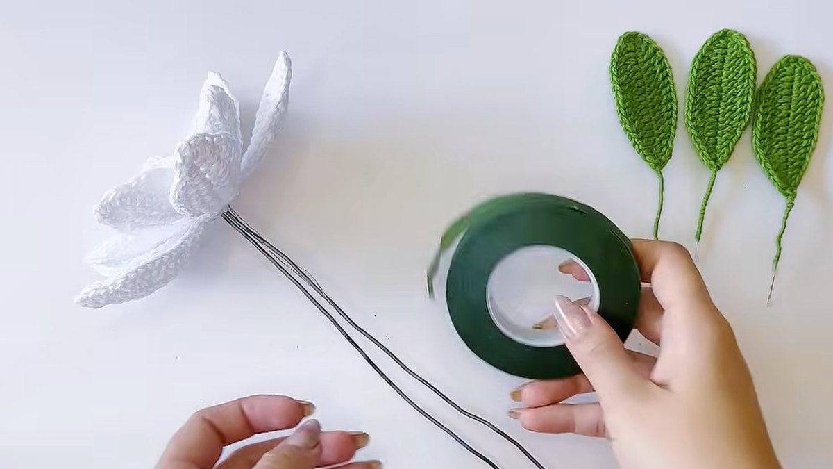 Green floral tape being wrapped round the stem wire of the crochet magnolia flower with three green leaves on the table